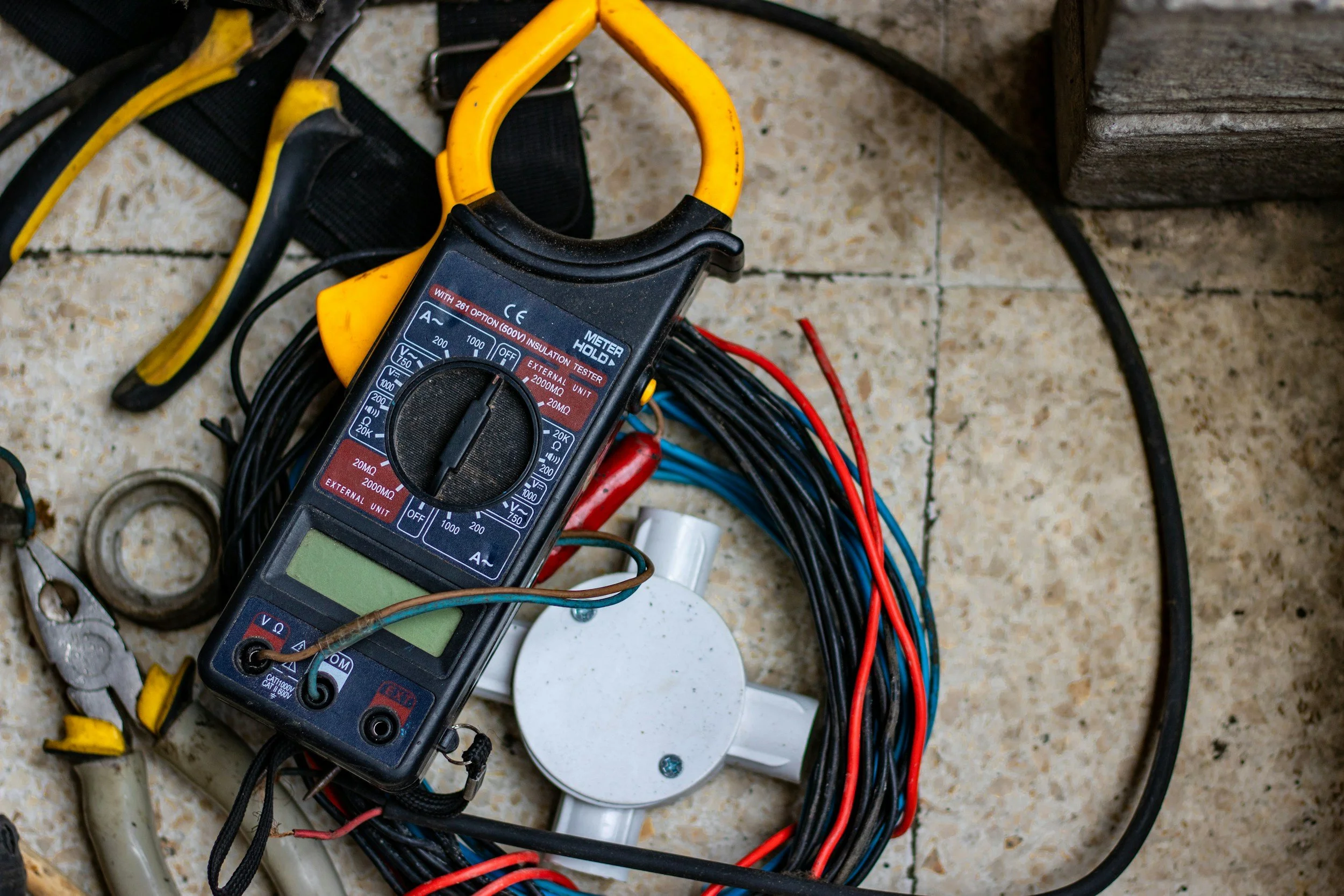 Digital multimeter surrounded by electrical tools and wires on a tiled floor.